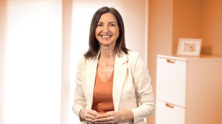 Fotografía de la psicóloga Julia Vidal sonriendo de forma cercana y profesional en el interior de su consulta. Viste una chaqueta blanca sobre una blusa cálida, en un entorno de terapia acogedor y luminoso. Imagen representativa de la sección «Desde la consulta».